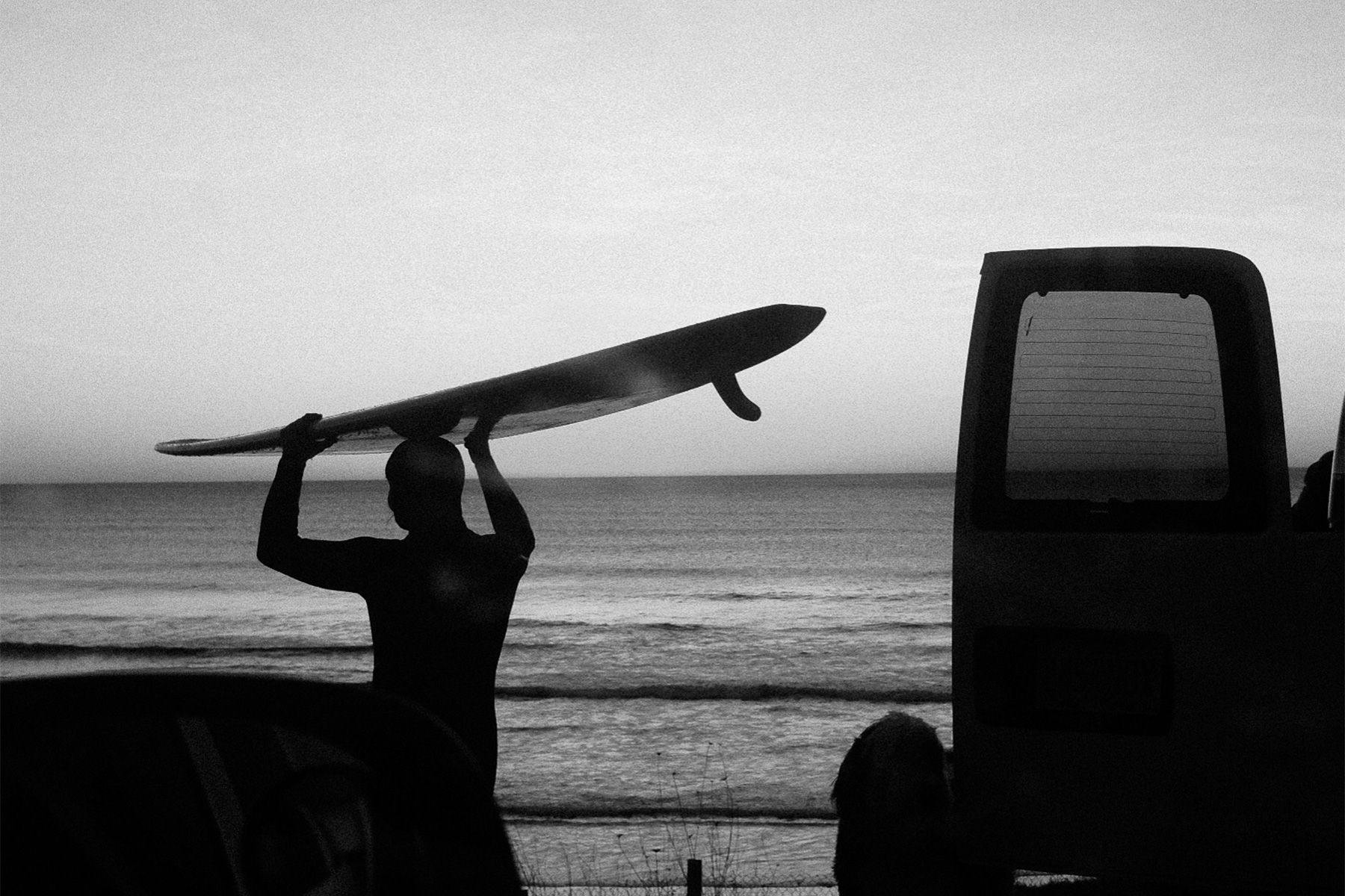 silhouette of a surfer carrying a longboard on their head past an open van door