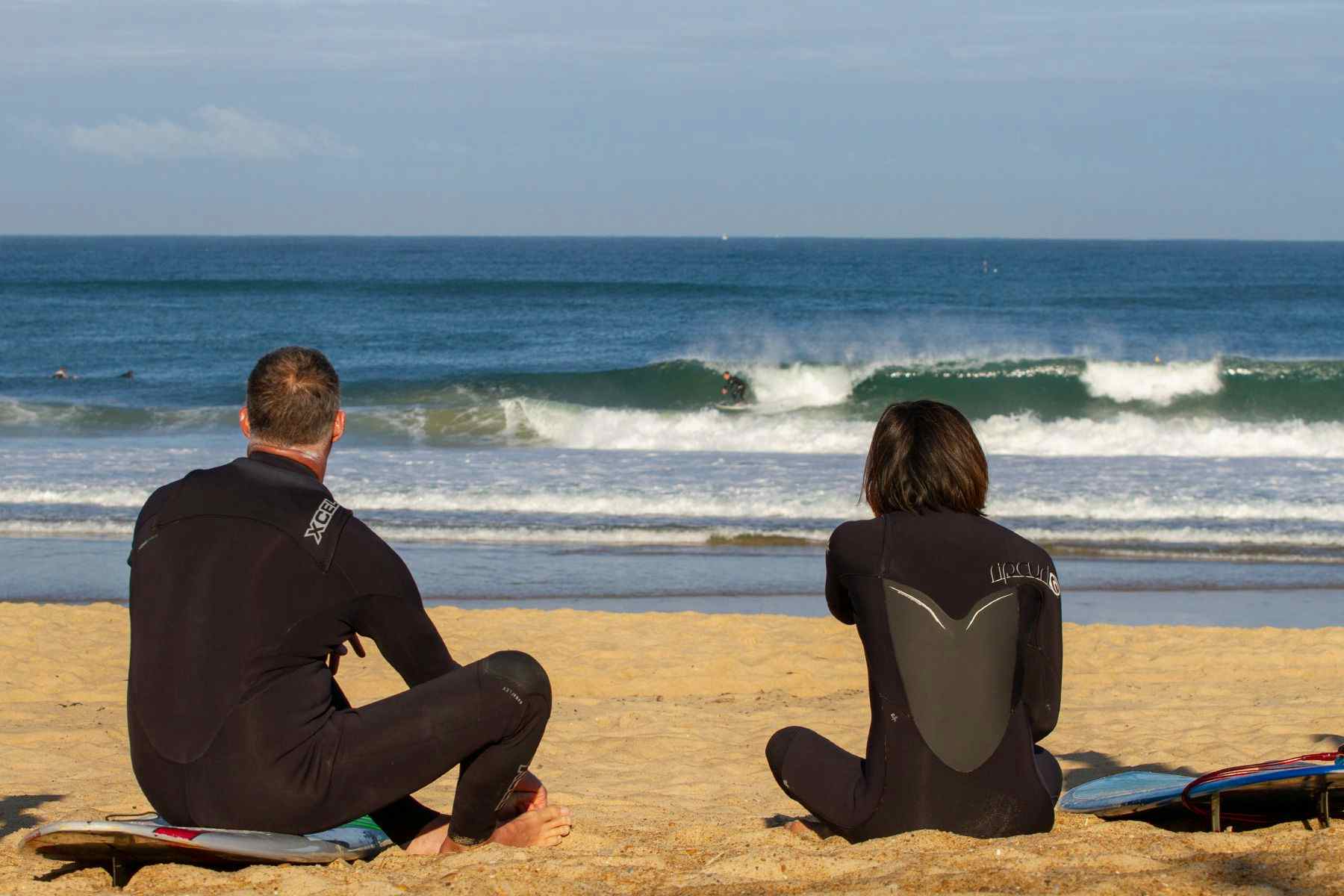 two surfers in wetsuits sitting on a sandy beach watching the surf