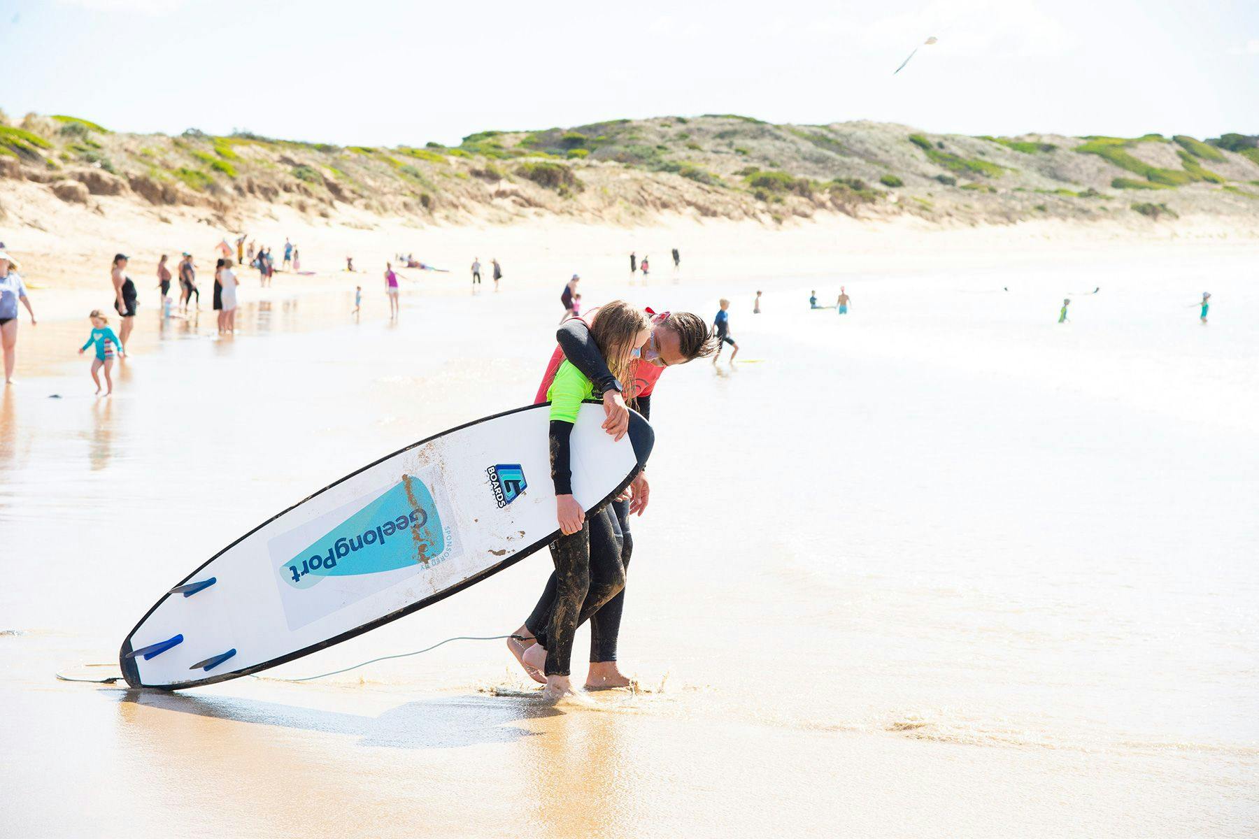 young person learning to surf and their surf coach carrying a surfboard to the ocean