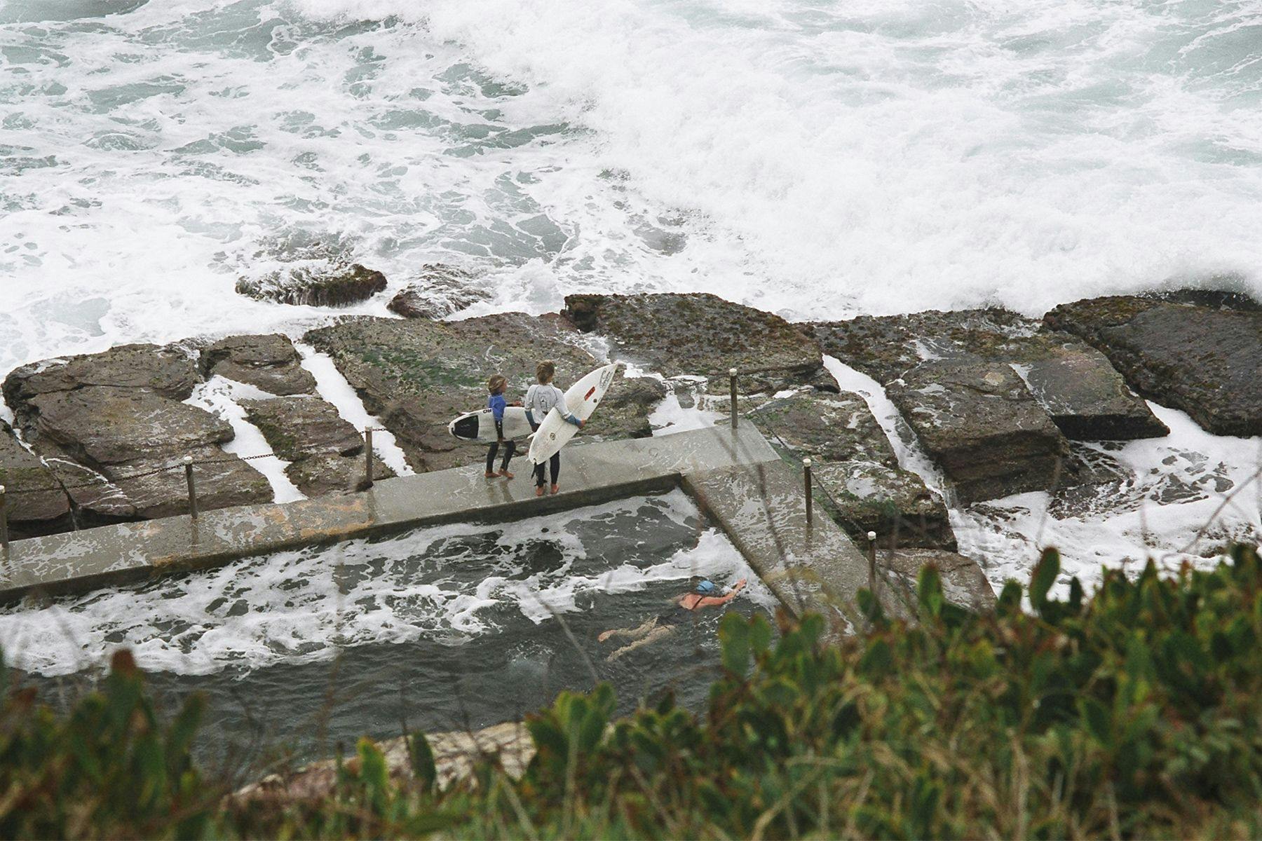 two young surfers wait by the edge of a tidal pool with a swimmer doing laps, ready to jump into the surf
