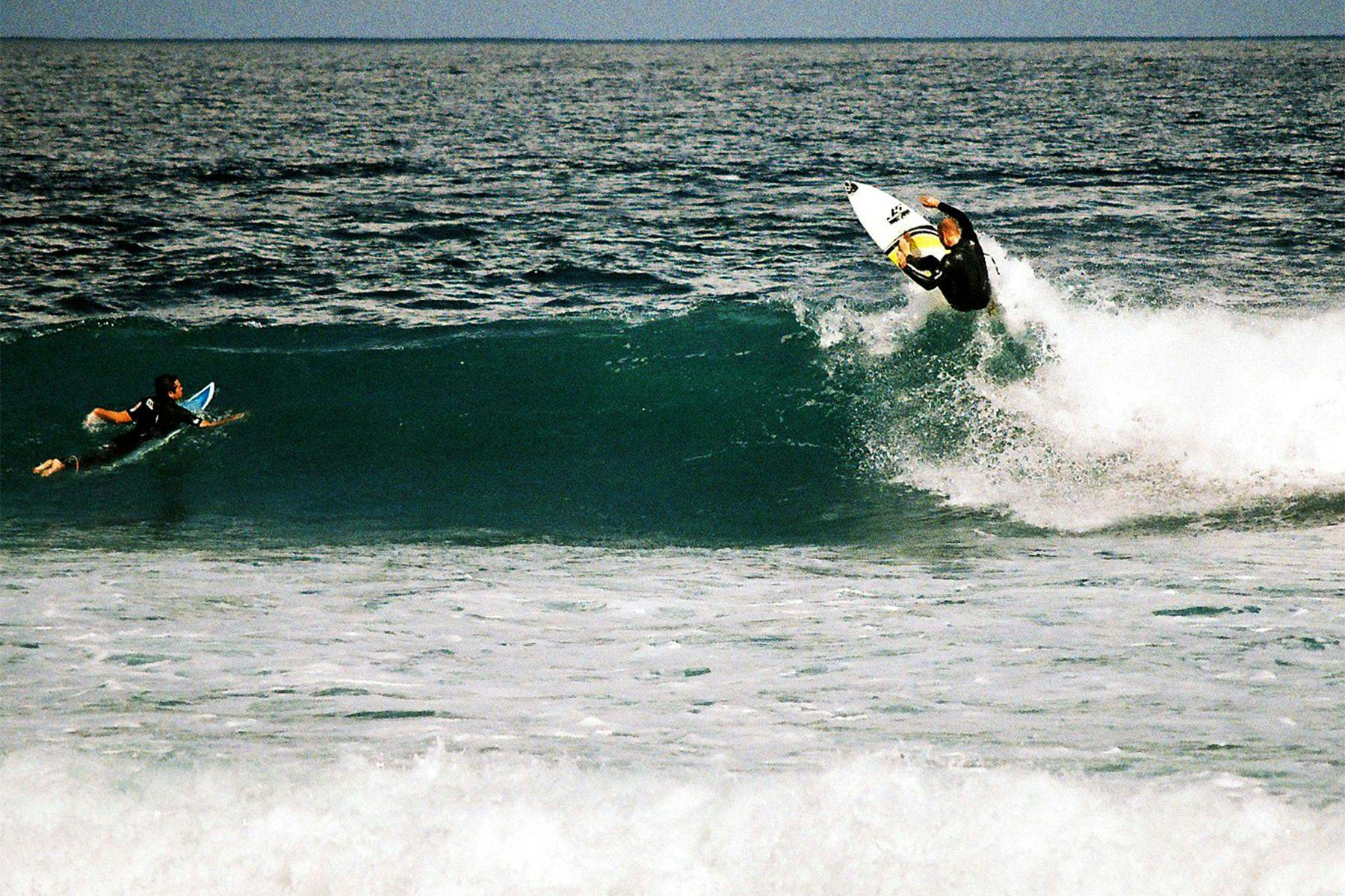 surfer doing a re-entry on a small right hand wave as another surfer paddles over the shoulder and watches 