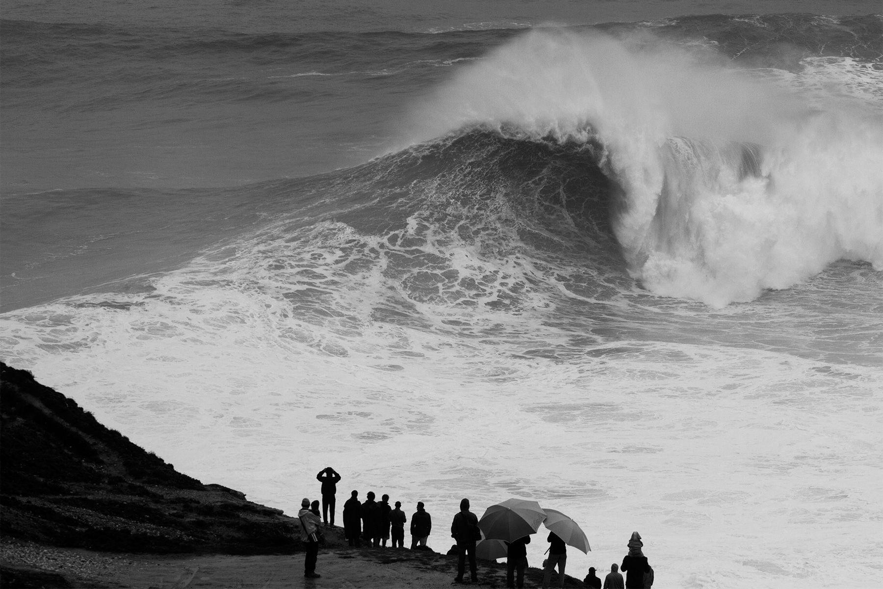 a group of people stood on the cliffs at nazare watching huge waves breaking