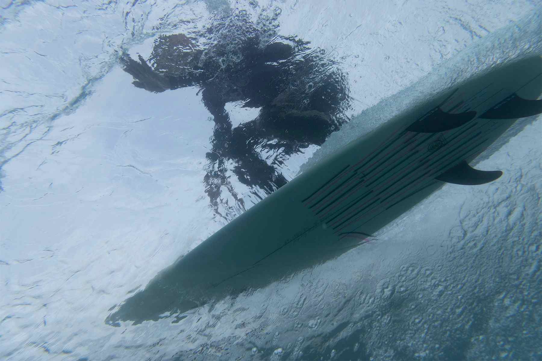 underwater view of a surfer going along a wave with tell tale ribbons on the bottom of his surfboard