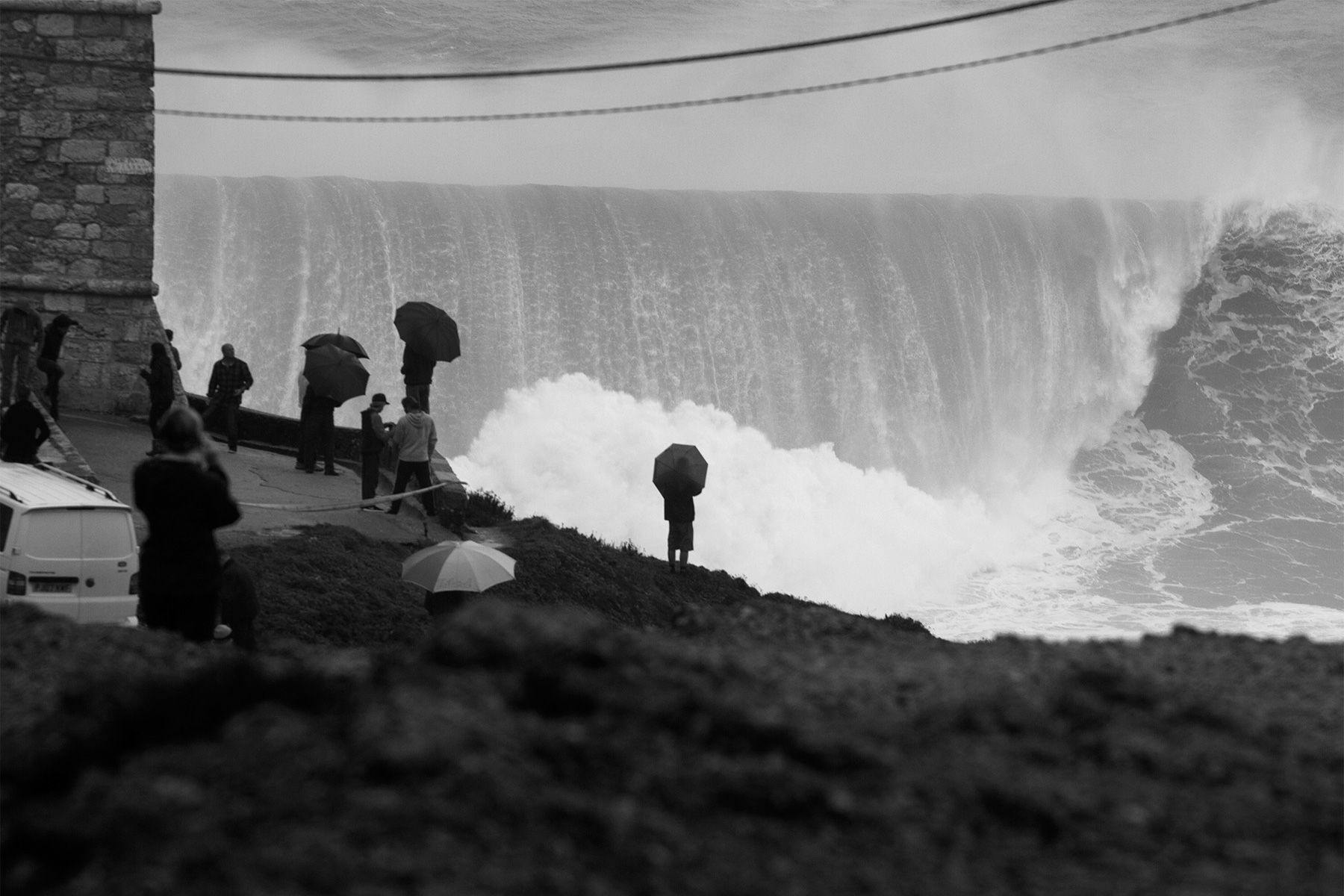 spectator with an umbrella watching a giant wave break at nazare in portugla